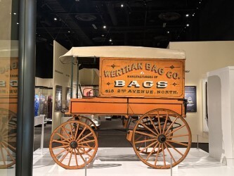 There is a brown covered wagon on display inside of a room at the museum.