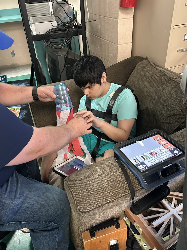 A student is sitting in a chair as a teacher helps them with their flag windsock.
