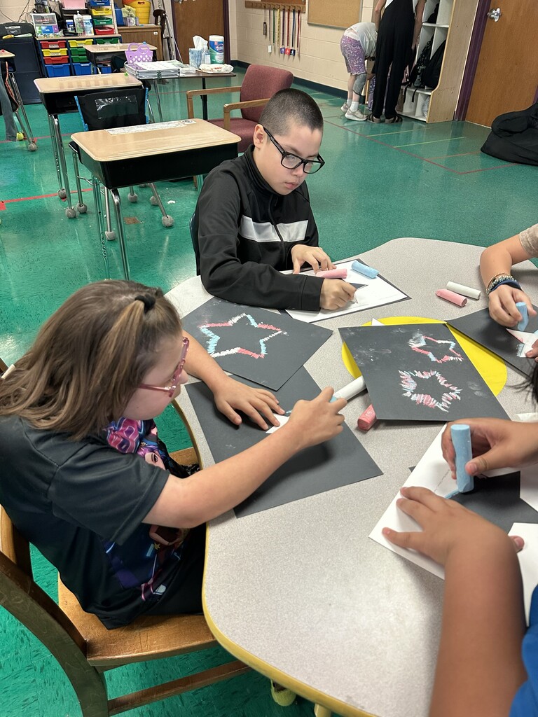Several students are seated at a table working on artwork that includes a star.
