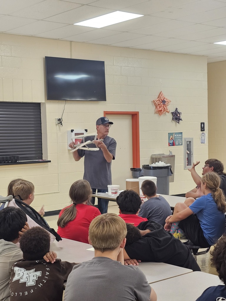 Mr. Shawn Clement from Nash Family Creamery holds up a piece of their ice cream machine during his presentation to students who are seated at cafeteria tables.