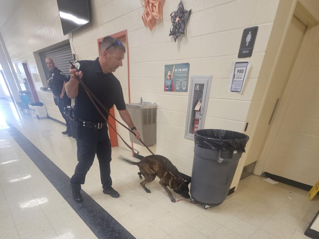 The officer has his K9 on a leash and is showing how the dog is sniffing a trash can to the students.