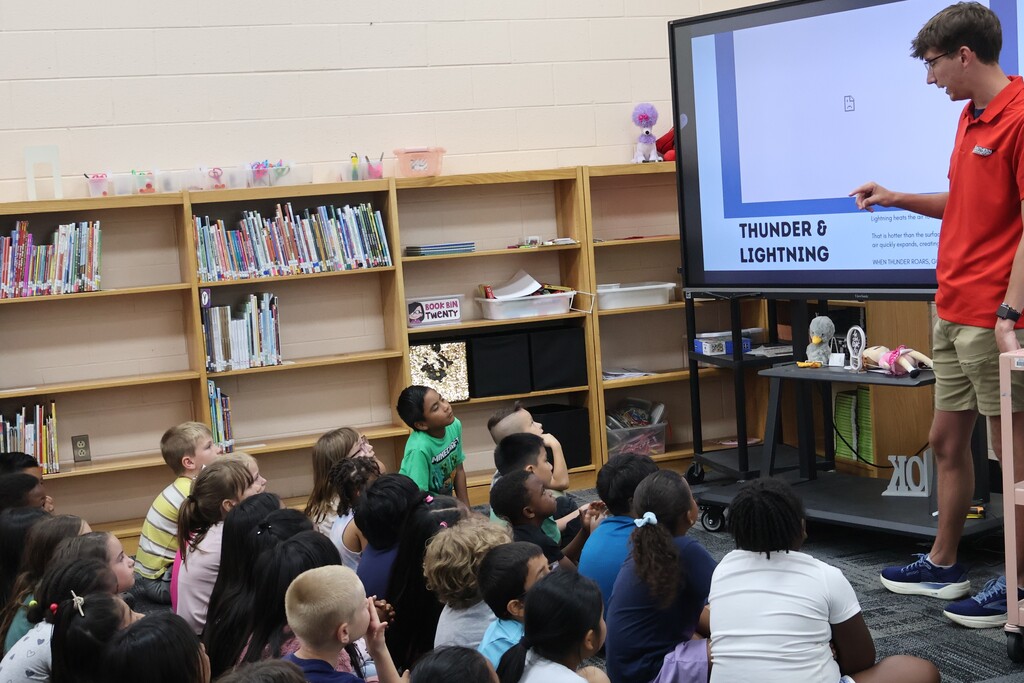 Students are seated on the carpet in front of  a large screen watching a presentation by Meteorologist Elijah Kirby about thunder & lightning. 