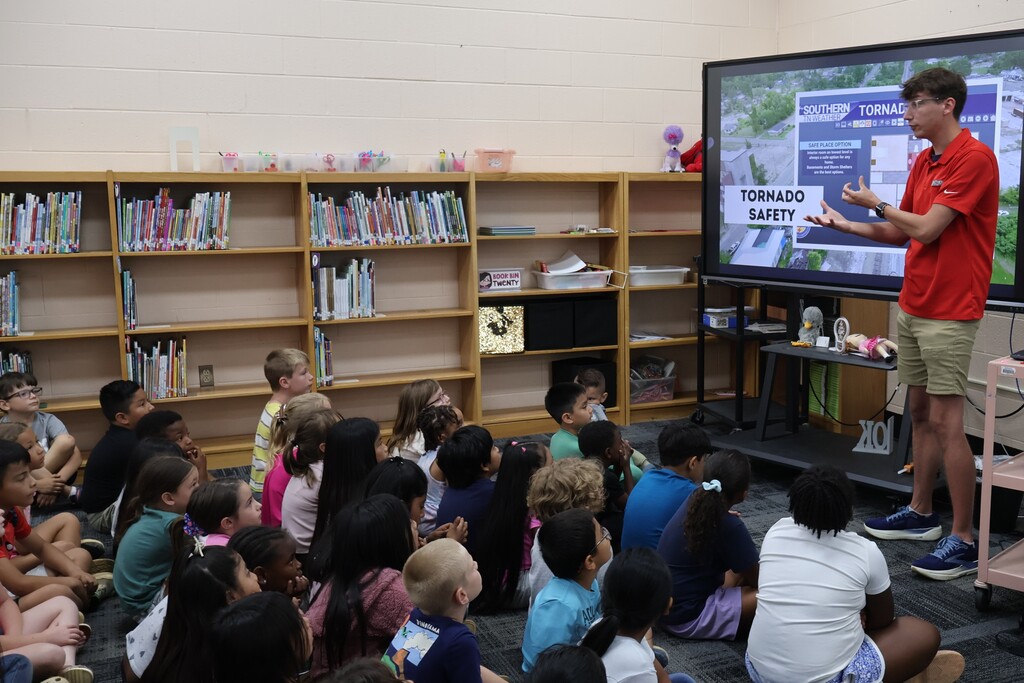 Students are seated on the carpet in front of  a large screen watching a presentation by Meteorologist Elijah Kirby about tornado safety.