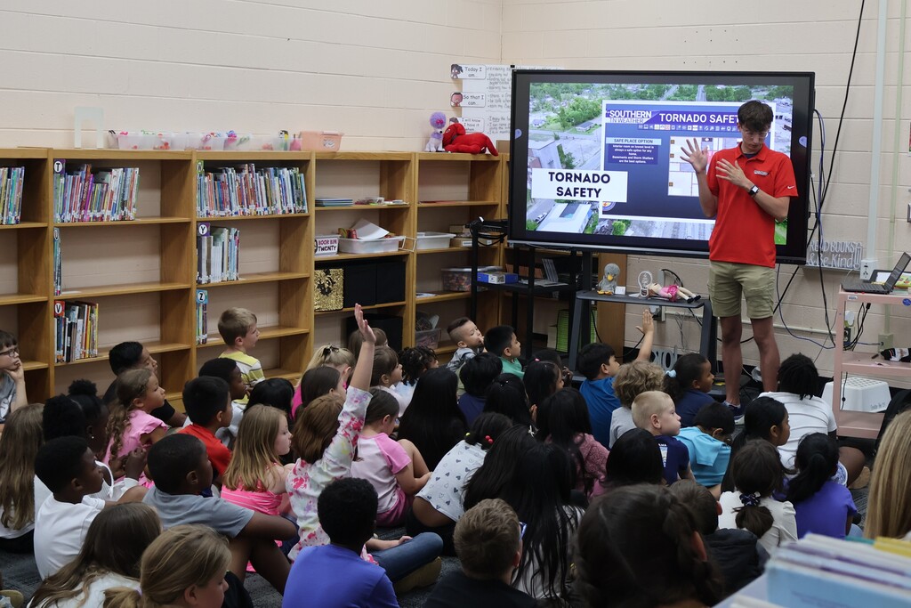 Students are seated on the carpet in front of  a large screen watching a presentation by Meteorologist Elijah Kirby about tornado safety.