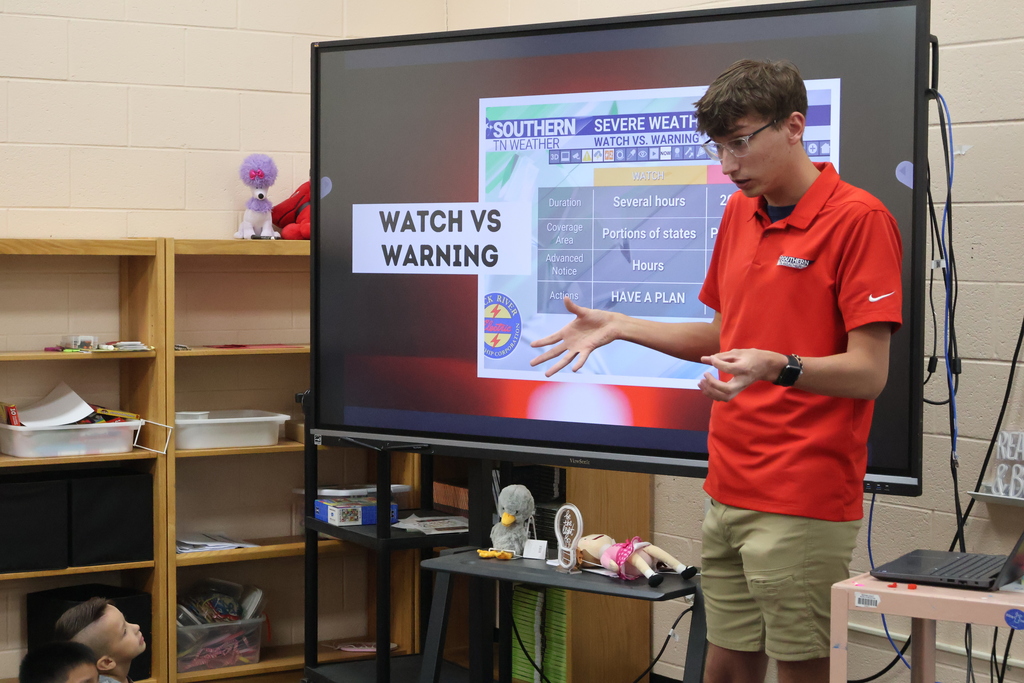 Students are seated on the carpet in front of  a large screen watching a presentation by Meteorologist Elijah Kirby about tornado safety.