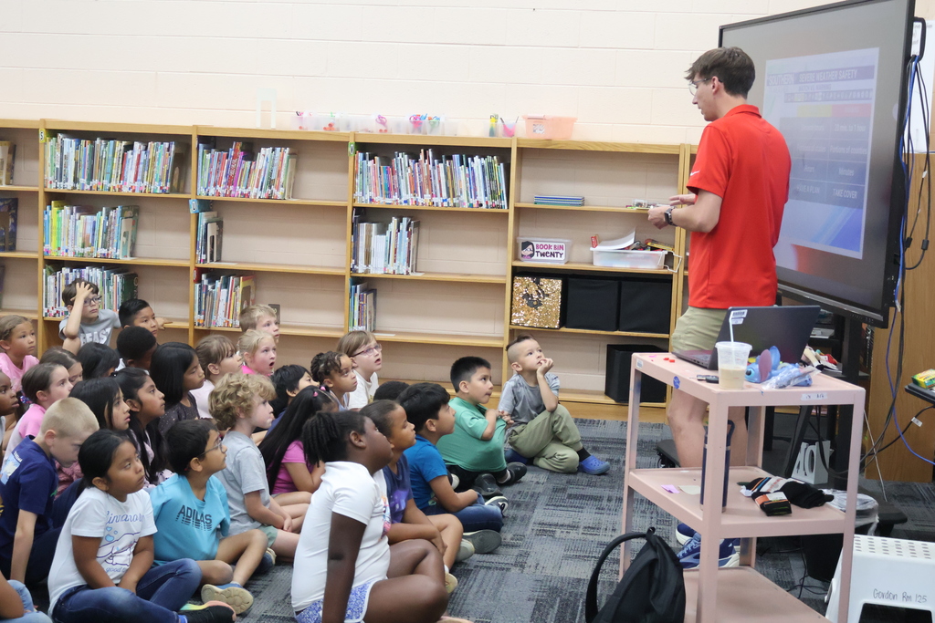 Students are seated on the carpet in front of  a large screen watching a presentation by Meteorologist Elijah Kirby about weather.