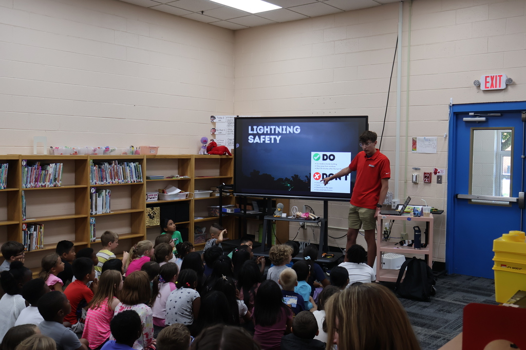 Students are seated on the carpet in front of  a large screen watching a presentation by Meteorologist Elijah Kirby about lightning safety. 