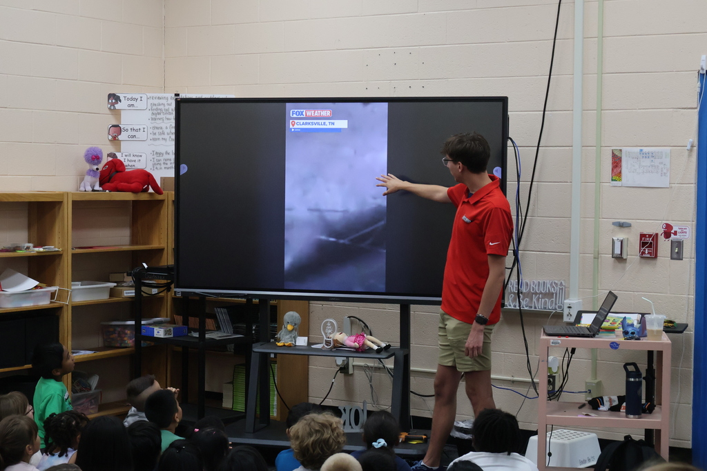 Students are seated on the carpet in front of  a large screen watching a presentation by Meteorologist Elijah Kirby about tornados. 