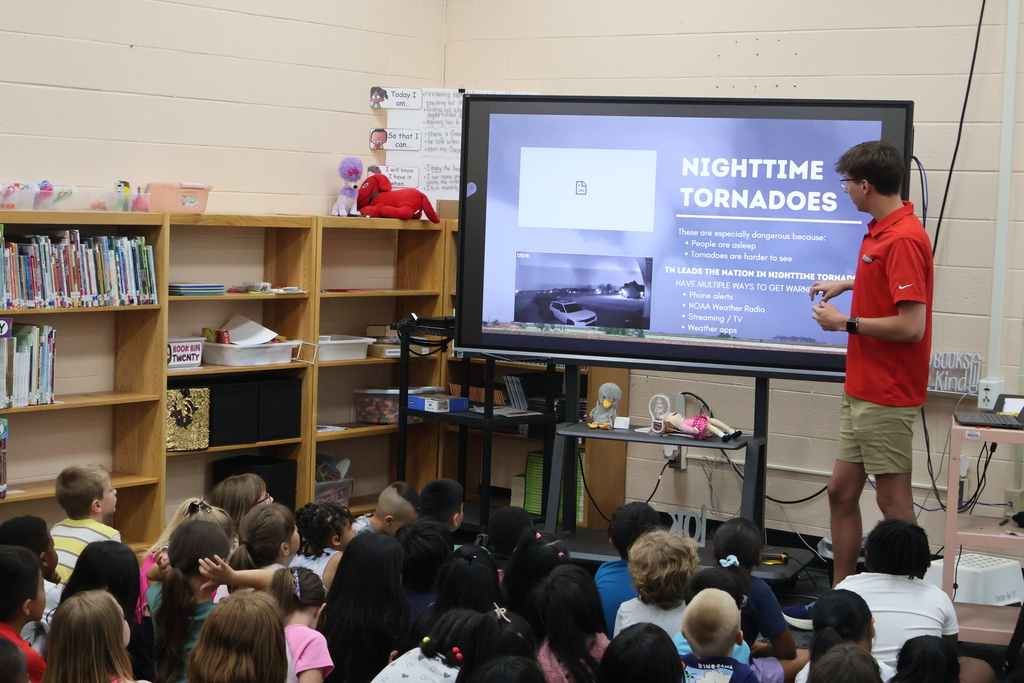 Students are seated on the carpet in front of  a large screen watching a presentation by Meteorologist Elijah Kirby about nighttime tornados.