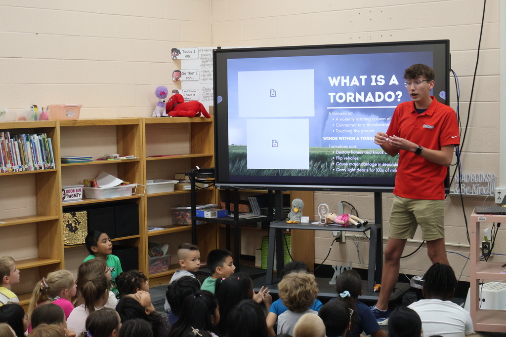 Students are seated on the carpet in front of  a large screen watching a presentation by Meteorologist Elijah Kirby about tornados. 