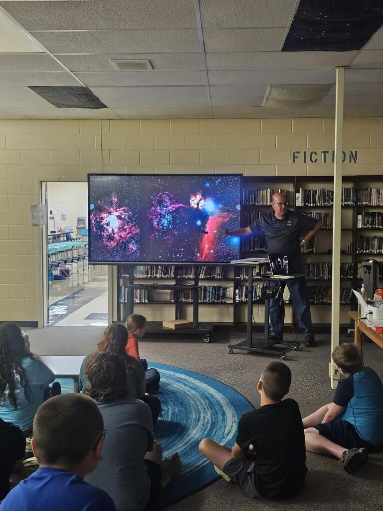 Mr. Billy Hix stands in the library beside a large screen as students are seated on the carpet watching his presentation on Astronomy.