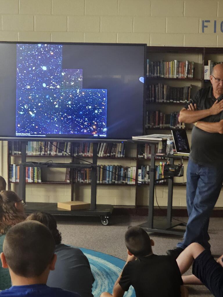 Mr. Billy Hix stands in the library beside a large screen as students are seated on the carpet watching his presentation on Astronomy.