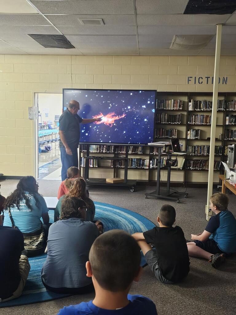Mr. Billy Hix stands in the library beside a large screen as students are seated on the carpet watching his presentation on Astronomy.
