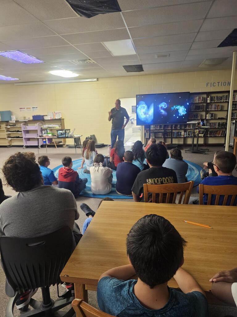 Mr. Billy Hix stands in the library beside a large screen as students are seated on the carpet watching his presentation on Astronomy.