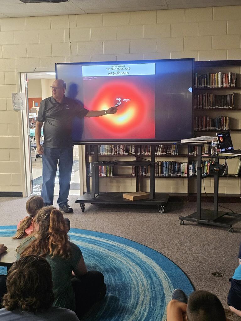 Mr. Billy Hix stands in the library beside a large screen as students are seated on the carpet watching his presentation on Astronomy.