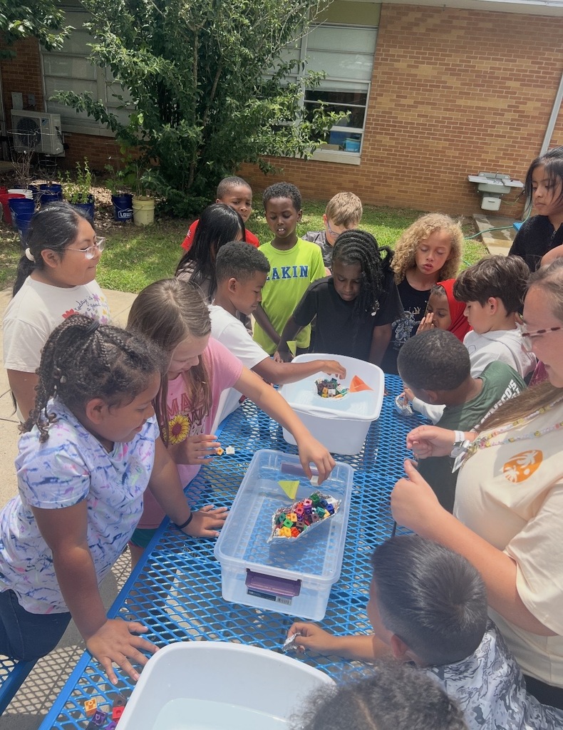 Students are standing outside around a table and a bucket of water placing cubes on their aluminum foil boat that they created to see if it will still float. 