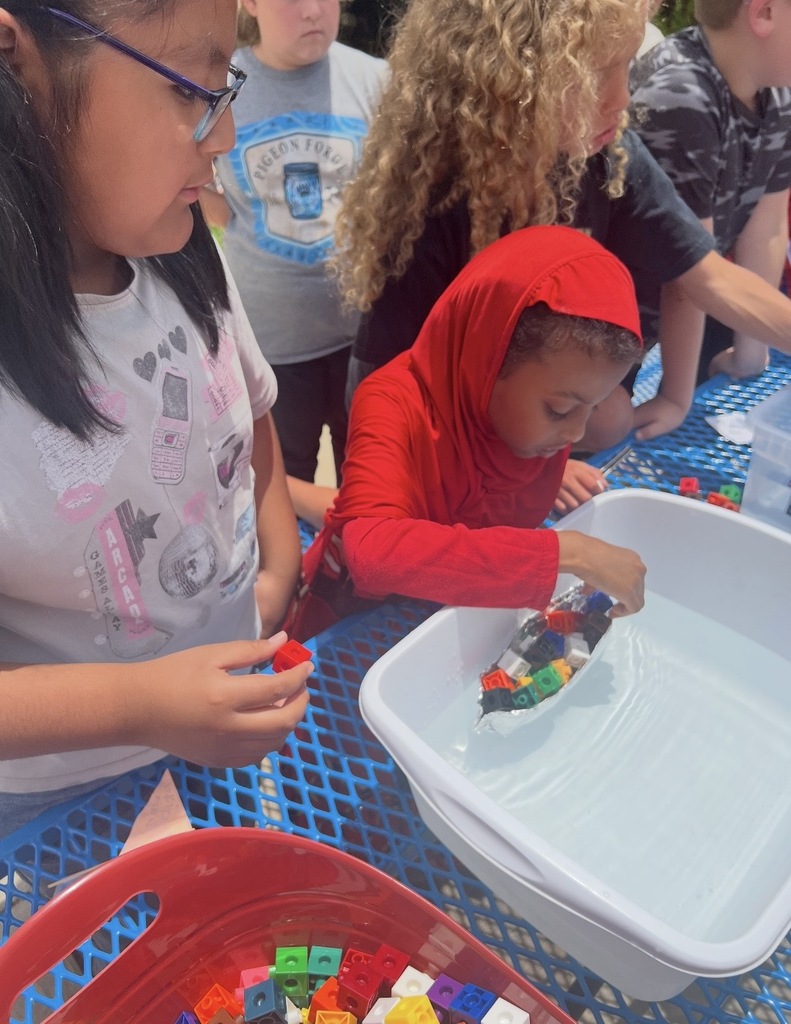 Students are standing outside around a table and a bucket of water placing cubes on their aluminum foil boat that they created to see if it will still float. 