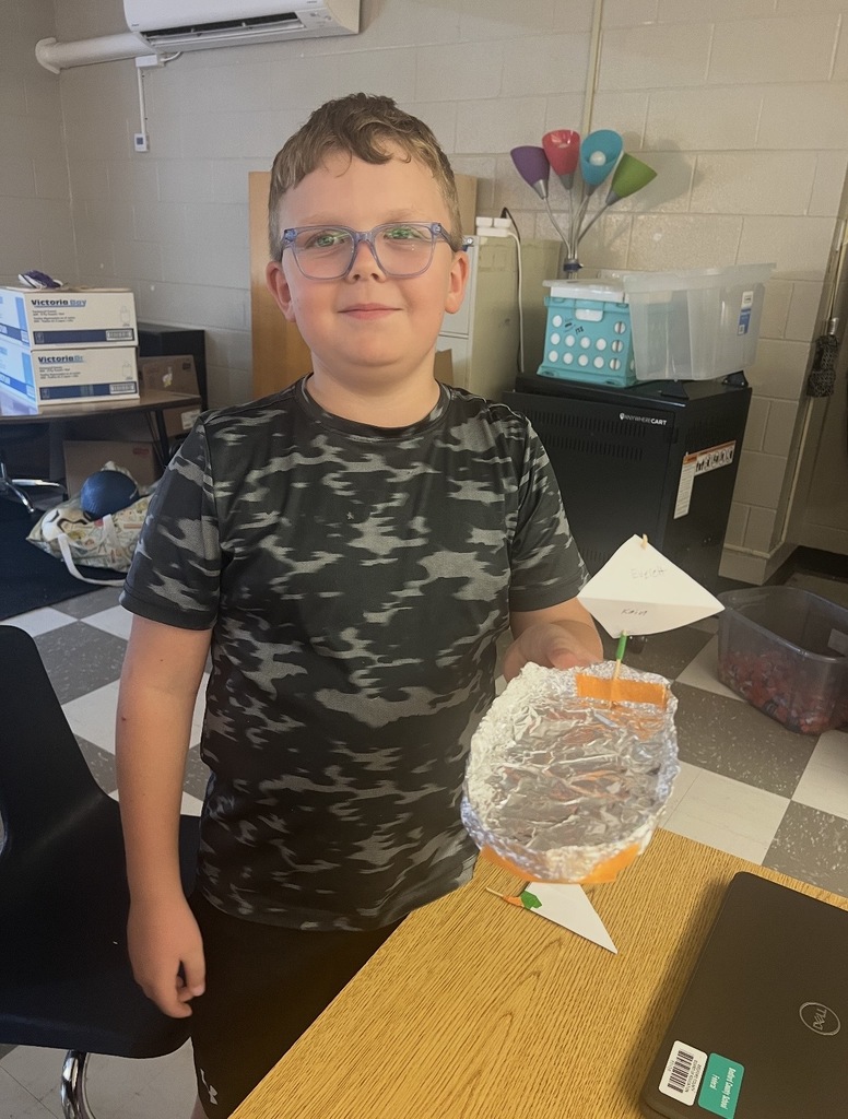 A student is standing at a desk holding up the boat he created with aluminum foil.