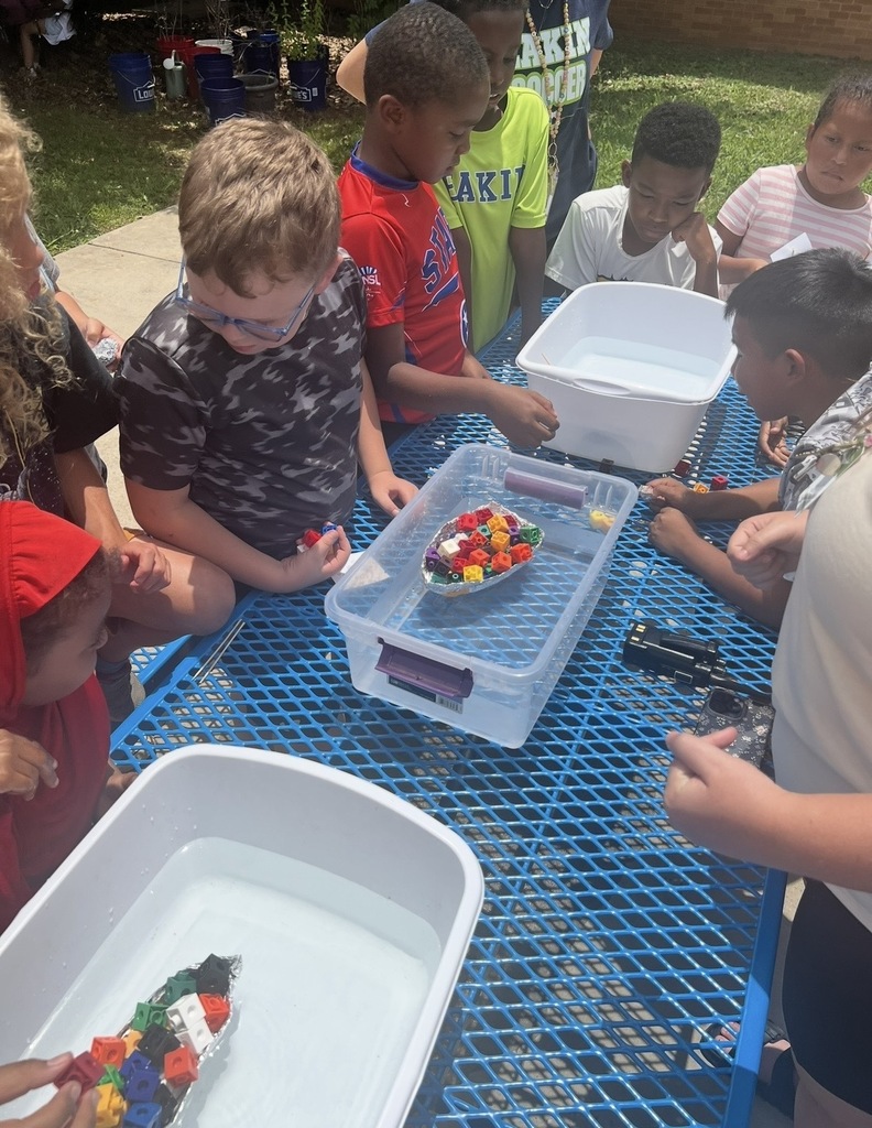 Students are standing outside around a table and a bucket of water placing cubes on their aluminum foil boat that they created to see if it will still float. 