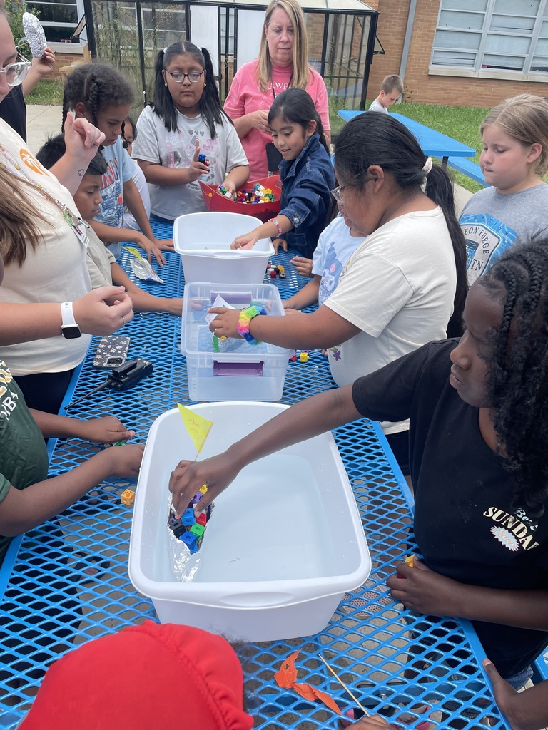 Students are standing outside around a table and a bucket of water placing cubes on their aluminum foil boat that they created to see if it will still float. 