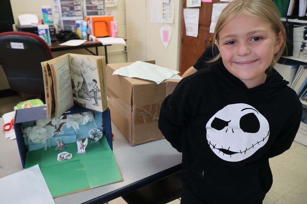 A student is sitting in a chair, holding a book in her hands, beside the diorama she created. 