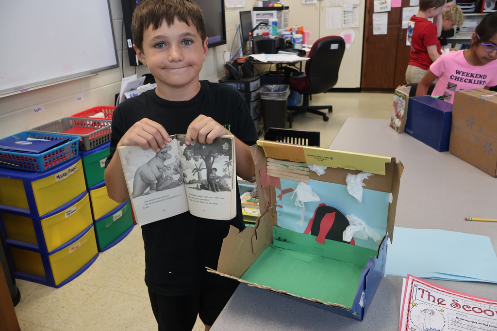 A student is standing beside a table holding a book in his hands, beside the diorama he created. 