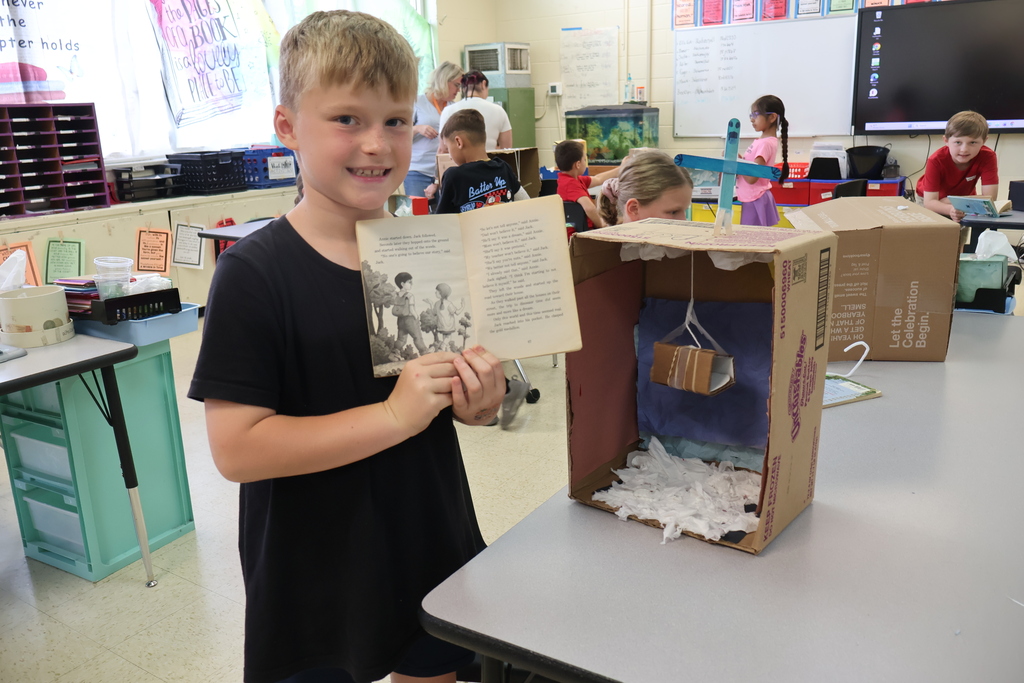 A student is standing beside a table holding a book in his hands, beside the diorama he created. 