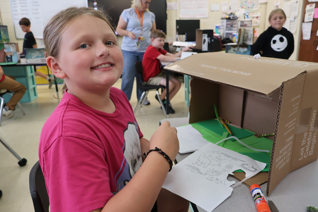 A student is sitting in a chair, holding a book in her hands, beside the diorama she created. 