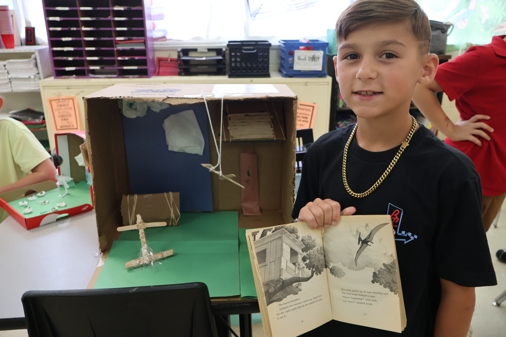 A student is standing beside a table holding a book in his hands, beside the diorama he created. 