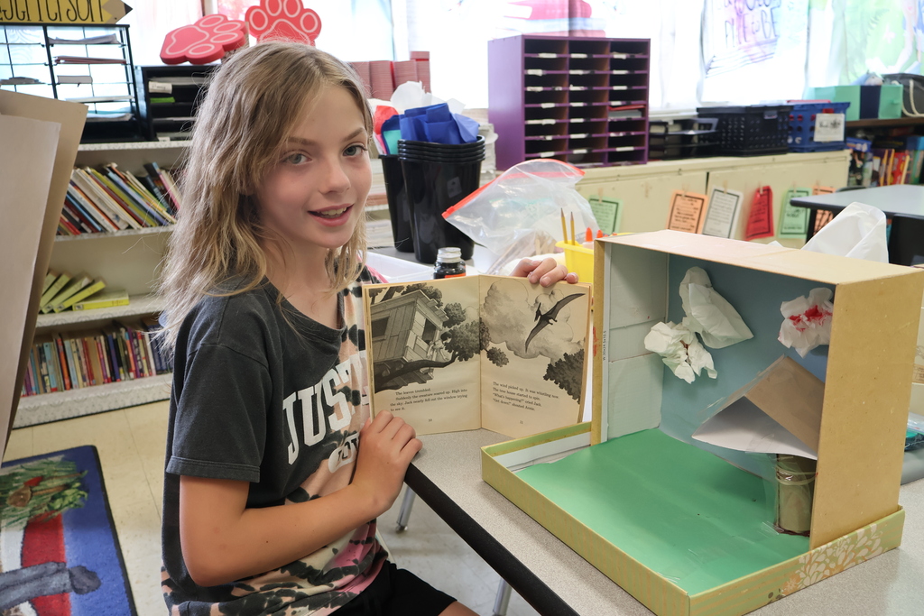 A student is sitting in a chair, holding a book in her hands, beside the diorama she created. 