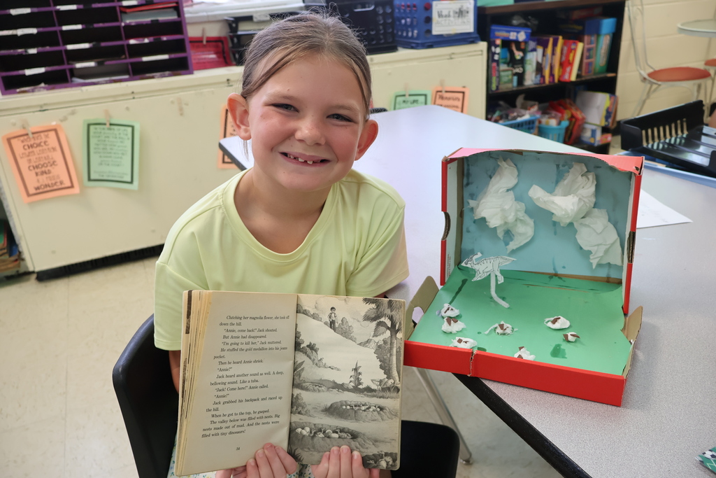A student is sitting in a chair, holding a book in her hands, beside the diorama she created. 