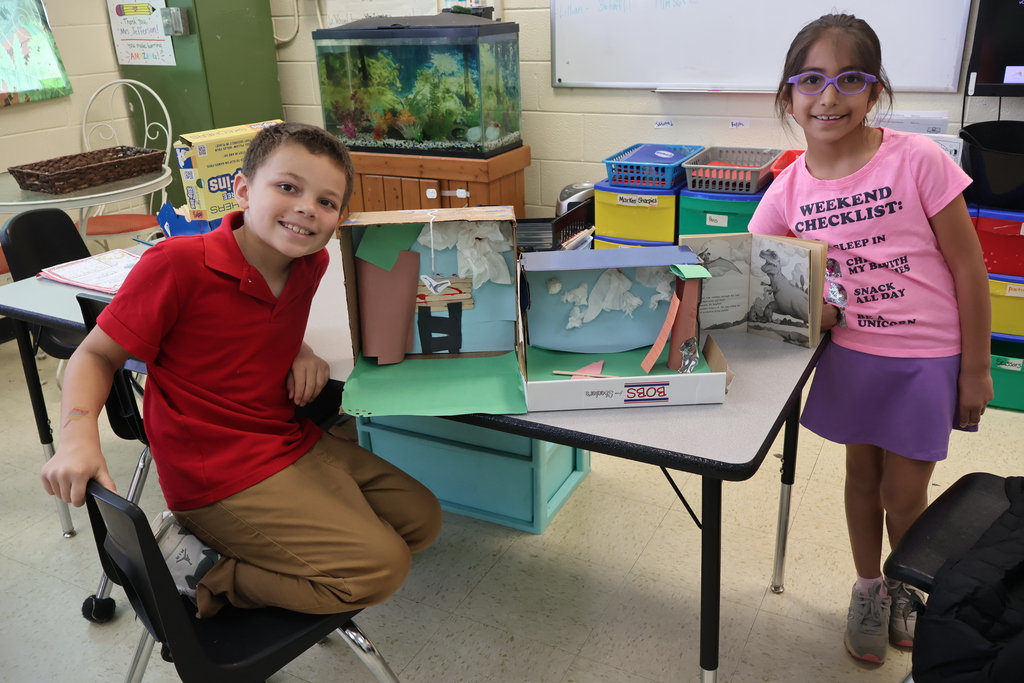Two students are sitting in a chair beside the dioramas they created. 