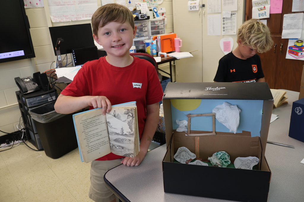 A student is standing beside a table holding a book in his hands, beside the diorama he created. 