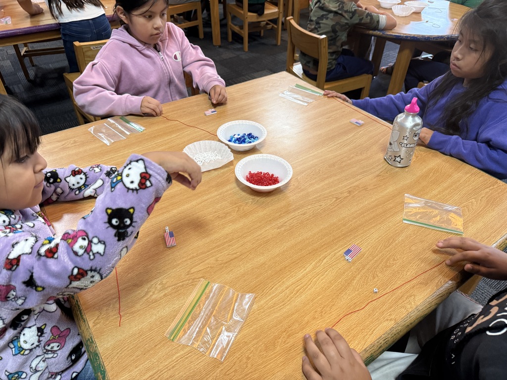 There are several tables with students seated at each making bracelets out of red, white, and blue beads.
