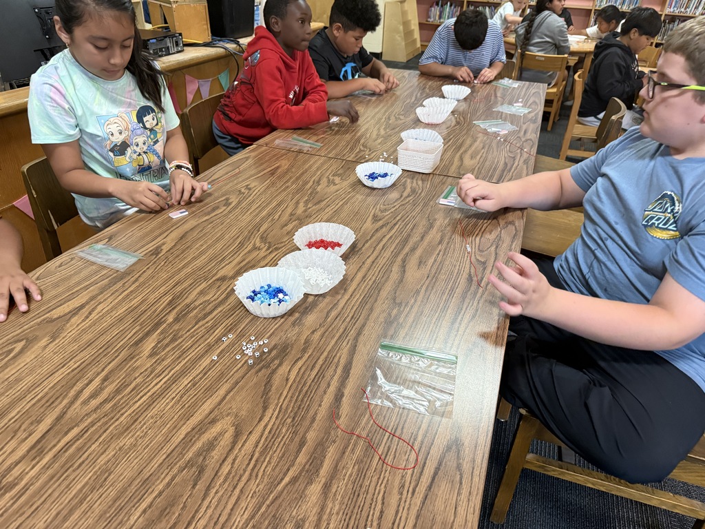 There are several tables with students seated at each making bracelets out of red, white, and blue beads.
