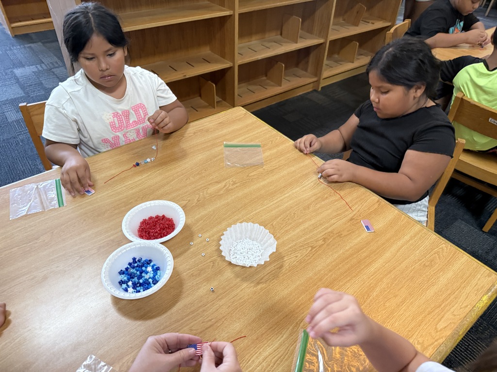 There are several tables with students seated at each making bracelets out of red, white, and blue beads.