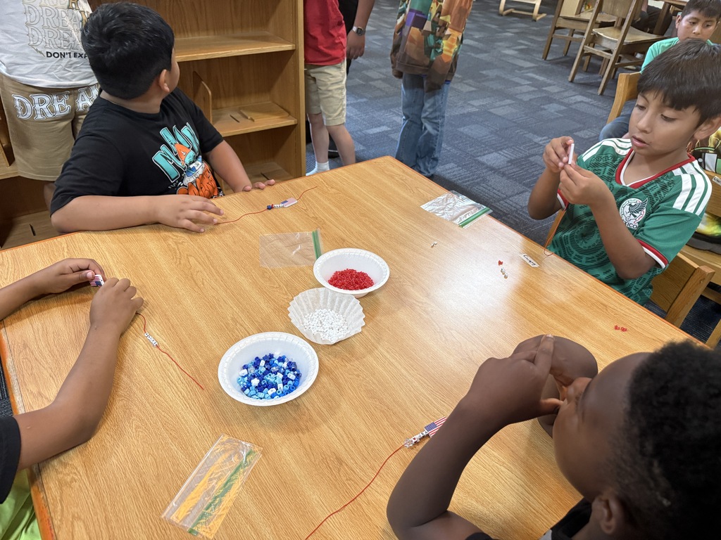 There are several tables with students seated at each making bracelets out of red, white, and blue beads.