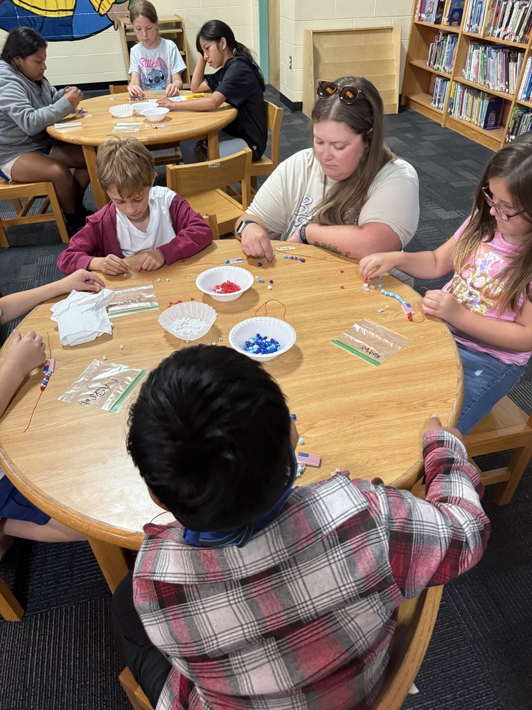 There are several tables with students seated at each making bracelets out of red, white, and blue beads.