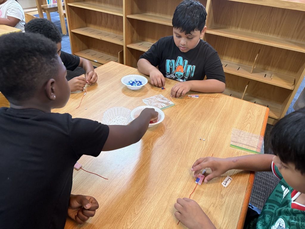 There are several tables with students seated at each making bracelets out of red, white, and blue beads.