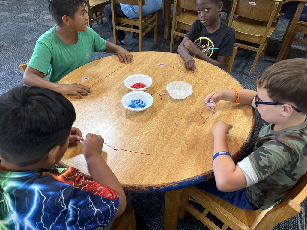 There are several tables with students seated at each making bracelets out of red, white, and blue beads.