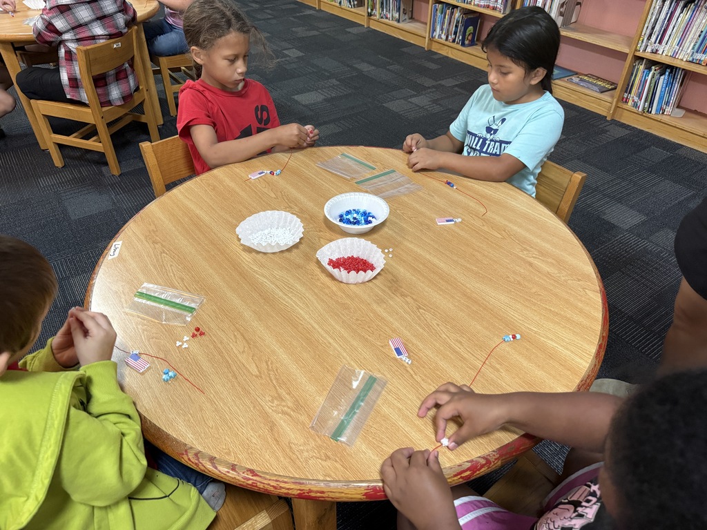 There are several tables with students seated at each making bracelets out of red, white, and blue beads.