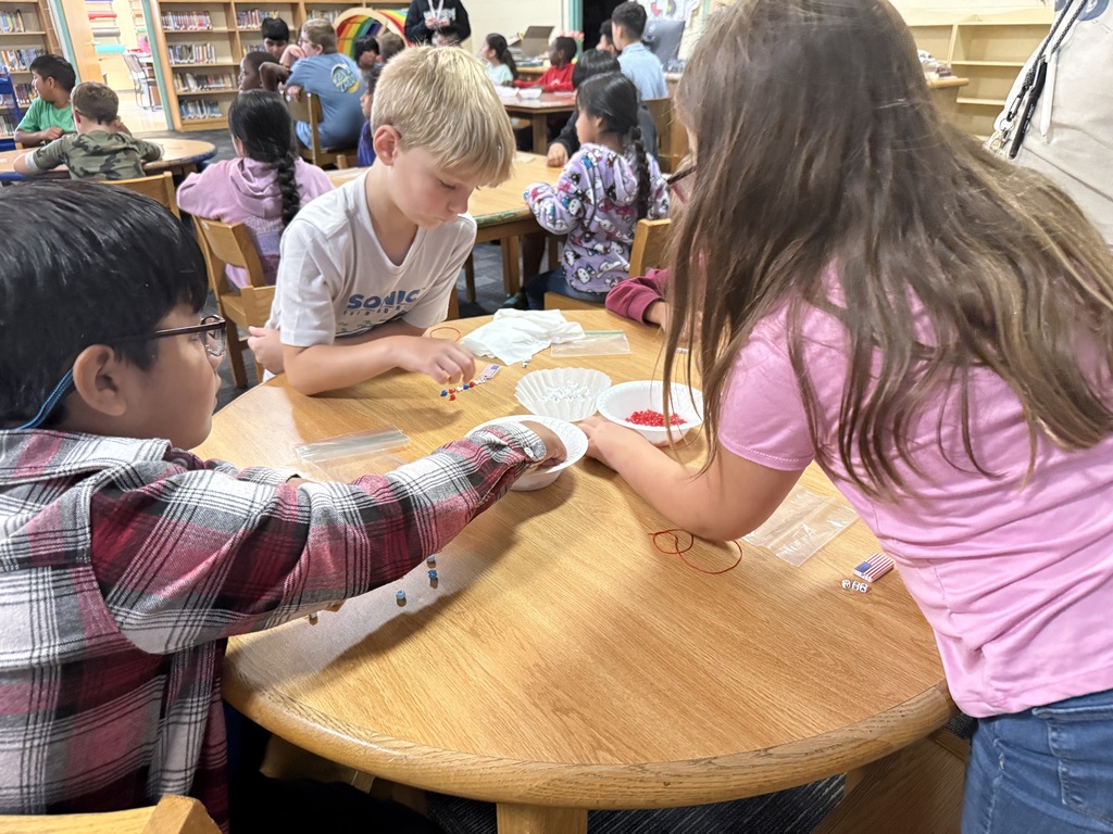 There are several tables with students seated at each making bracelets out of red, white, and blue beads.