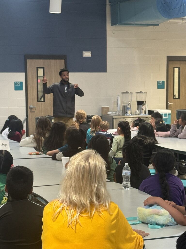 Jarious Wallce from Grand Canyon University speaks to teachers and students who are all seated at tables in the cafeteria.