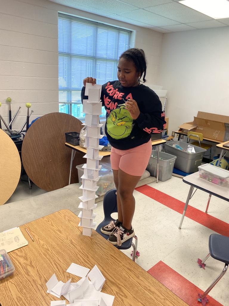 A student is standing on a chair beside her desk building a tower out of index cards.