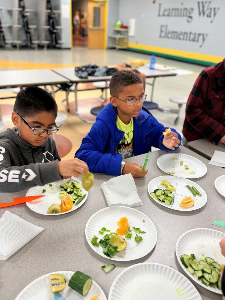 Two students are sitting down at a table with plates of fruits and vegetables for them to try. 