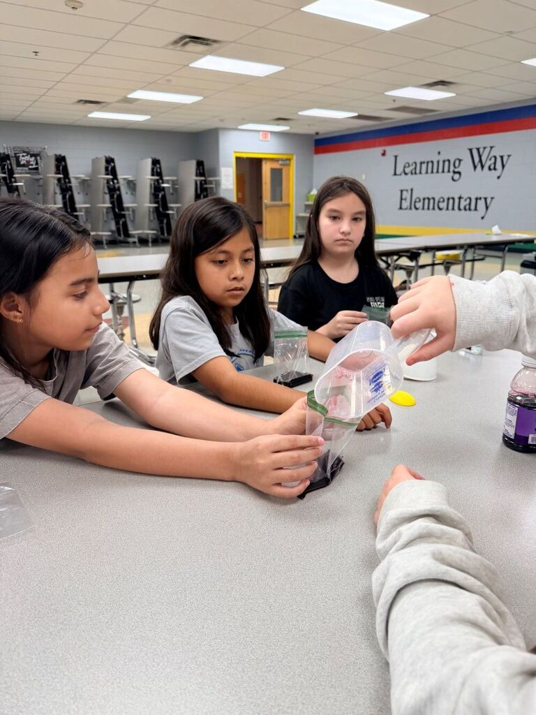 Three students are seated at a table pouring liquid into cups.