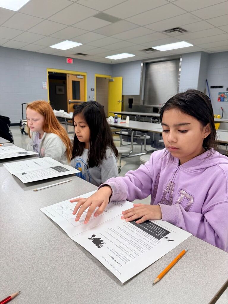 Three students are seated at a table reading over materials that were handed out. 
