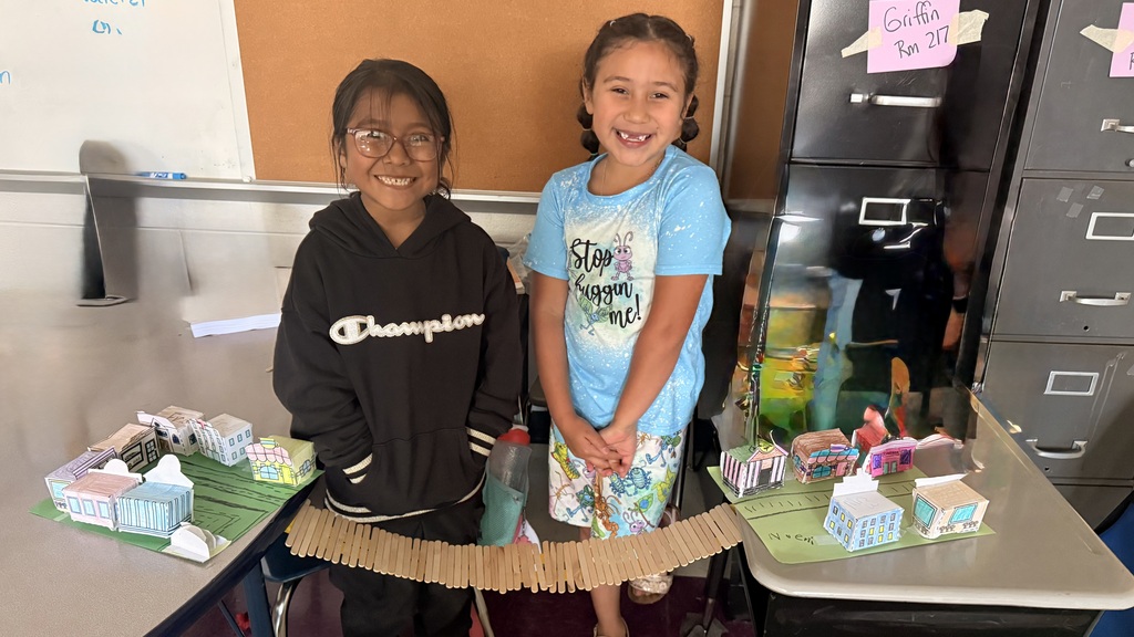 Two students are standing between two tables with the town and bridge they built.