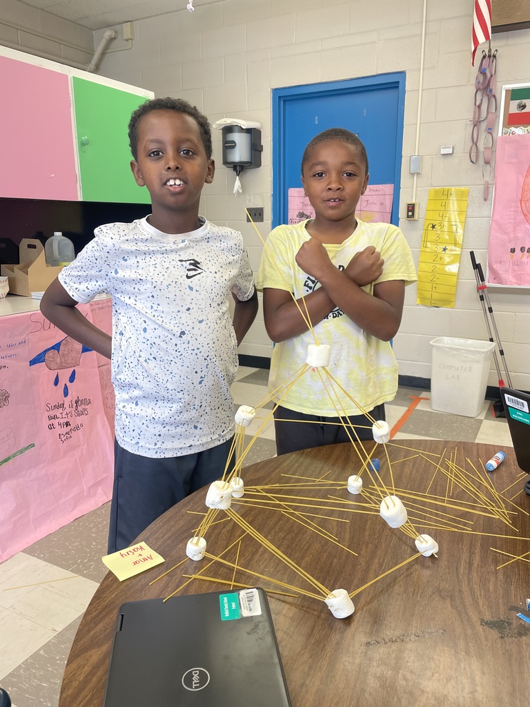 Two students are standing a table with their tower made from spaghetti and marshmallows.