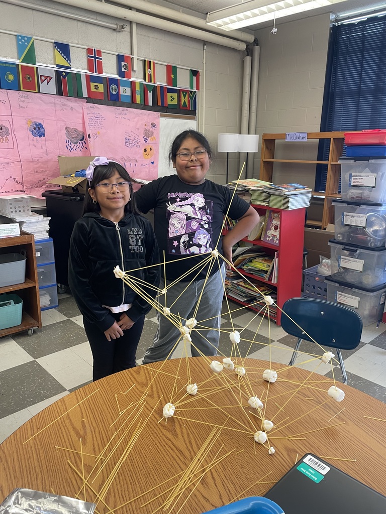 Two girls are standing a table with their tower made from spaghetti and marshmallows.